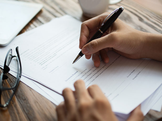 Unrecognizable businessman examining papers at table. Manager with ballpoint pen filling business papers. Close-up of male hands working at desk. Analyzing documents concept